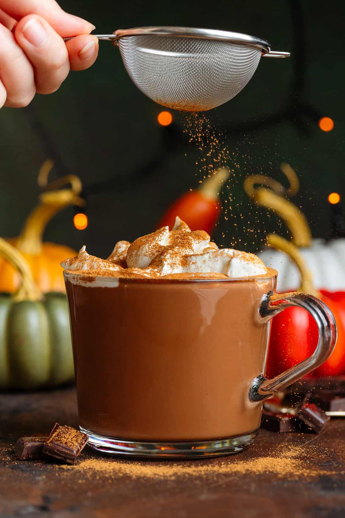 Creamy hot chocolate in a glass mug topped with homemade whipped cream being dusted with pumpkin pie spice with pumpkins in the background.