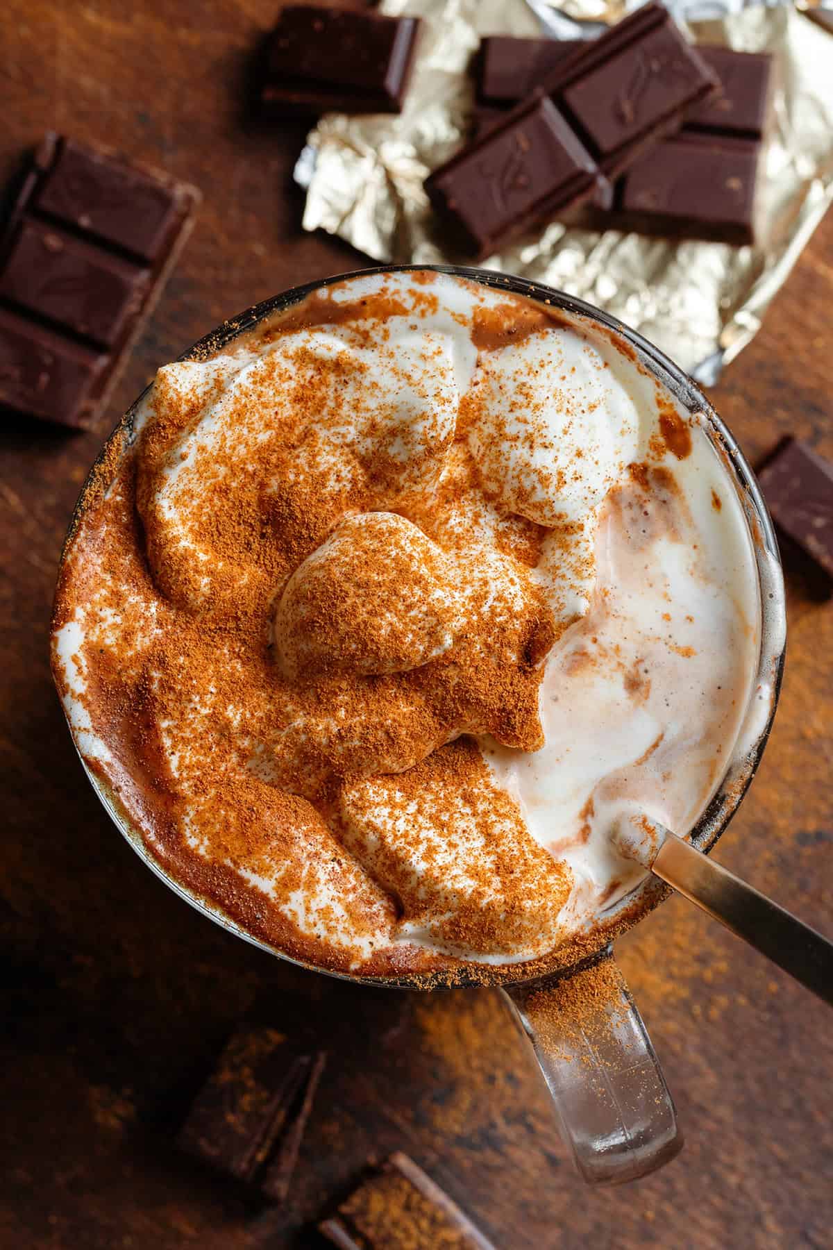 Creamy hot chocolate in a glass mug topped with whipped cream and dusted with pumpkin pie spice with some of the whipped cream eaten and a spoon resting inside the mug.