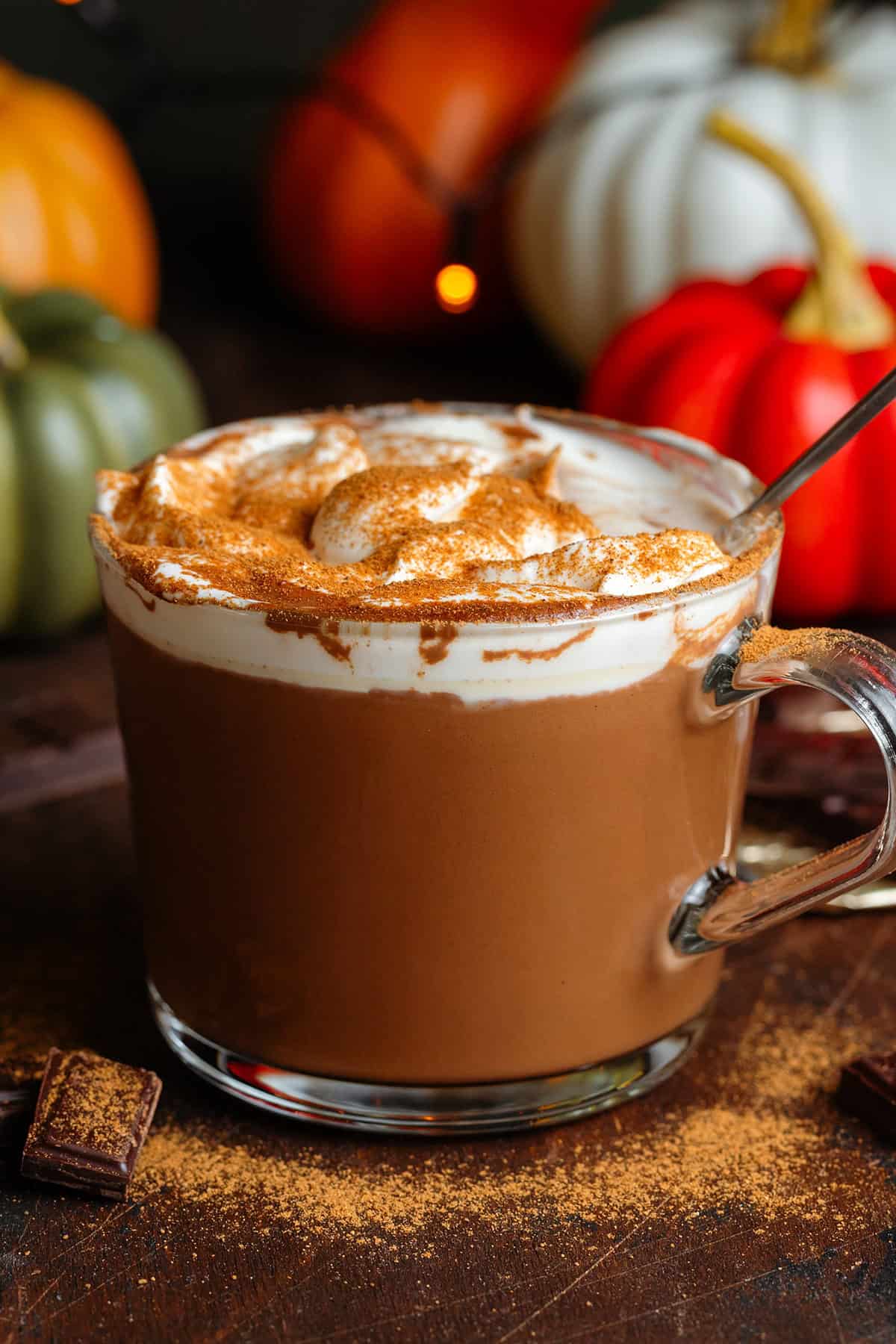 Creamy hot chocolate in a glass mug topped with whipped cream and dusted with pumpkin pie spice with a spoon in the mug after drinking a little bit of it.