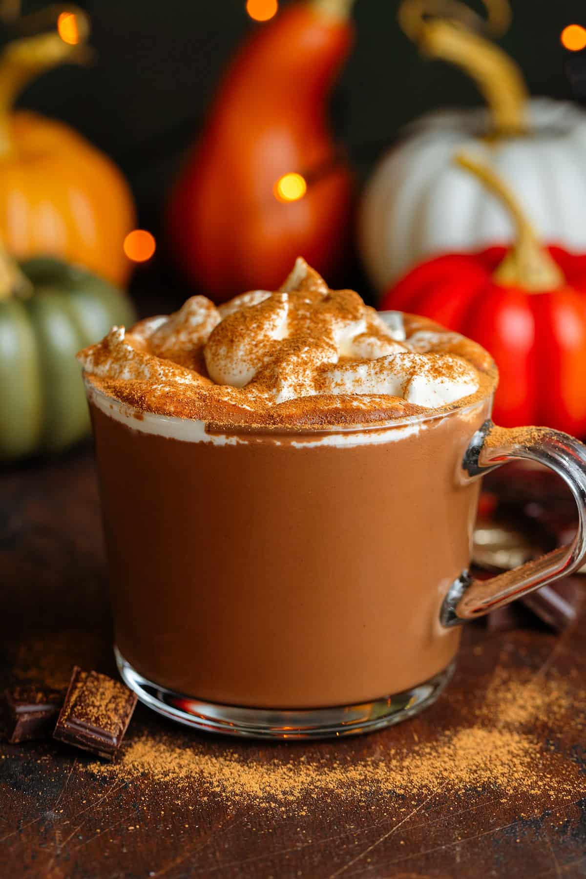 Creamy hot chocolate in a glass mug topped with whipped cream and dusted with pumpkin pie spice with chocolate squares around the mug.