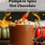 Creamy hot chocolate in a glass mug topped with whipped cream and being dusted with pumpkin pie spice.