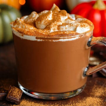 Creamy hot chocolate in a glass mug topped with whipped cream and dusted with pumpkin pie spice.