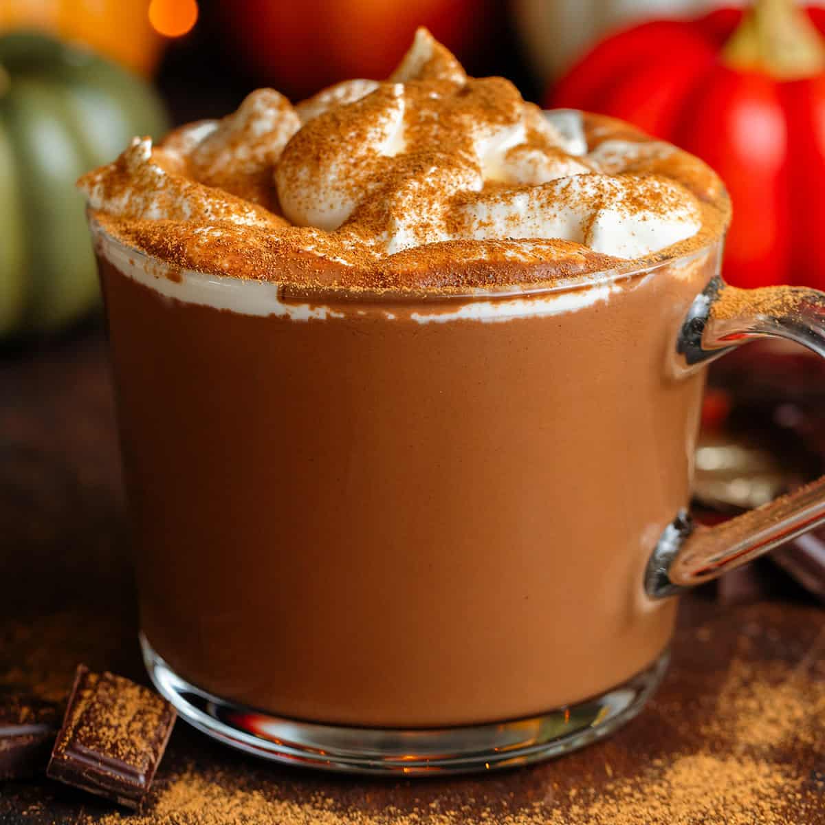Creamy hot chocolate in a glass mug topped with whipped cream and dusted with pumpkin pie spice.