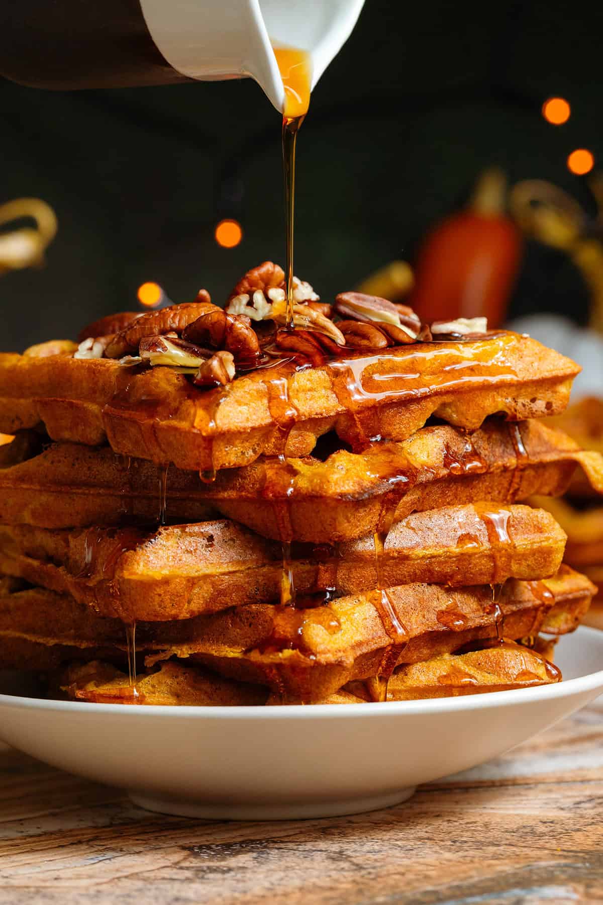 A stack of pumpkin pancakes on a small beige plate topped with chopped pecans being drizzled with maple syrup.