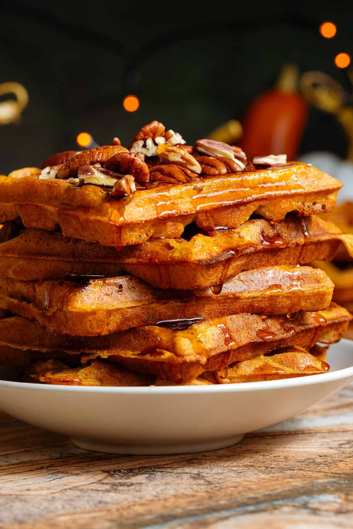 A stack of pumpkin pancakes topped with chopped pecans with maple syrup dripping down the sides on a small beige plate.