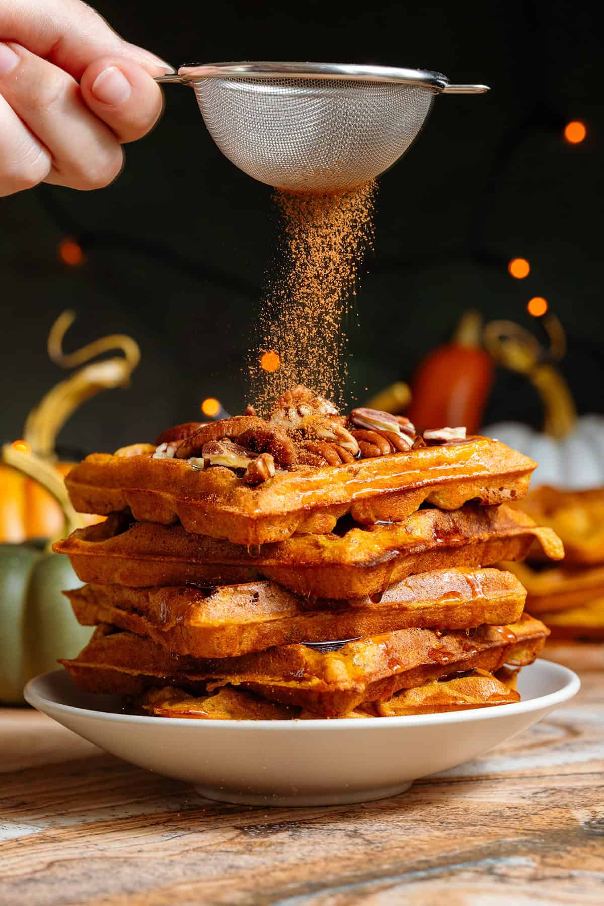 A stack of pumpkin pancakes topped with chopped pecans and drizzled with maple syrup on a small beige plate being dusted with pumpkin pie spice.
