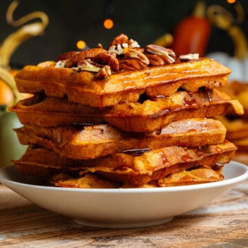 A stack of pumpkin pancakes on a small beige plate topped with chopped pecans with maple syrup dripping down the sides.