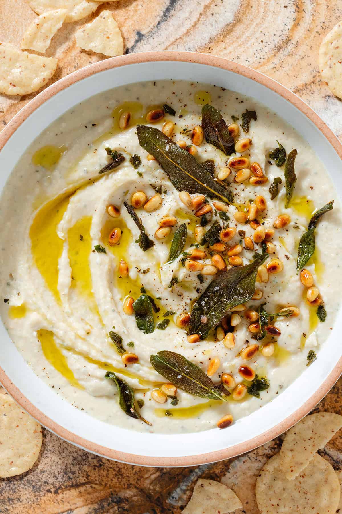 White bean dip in a low white bowl topped with fried sage leaves, toasted pine nuts, and olive oil with crackers around the bowl.