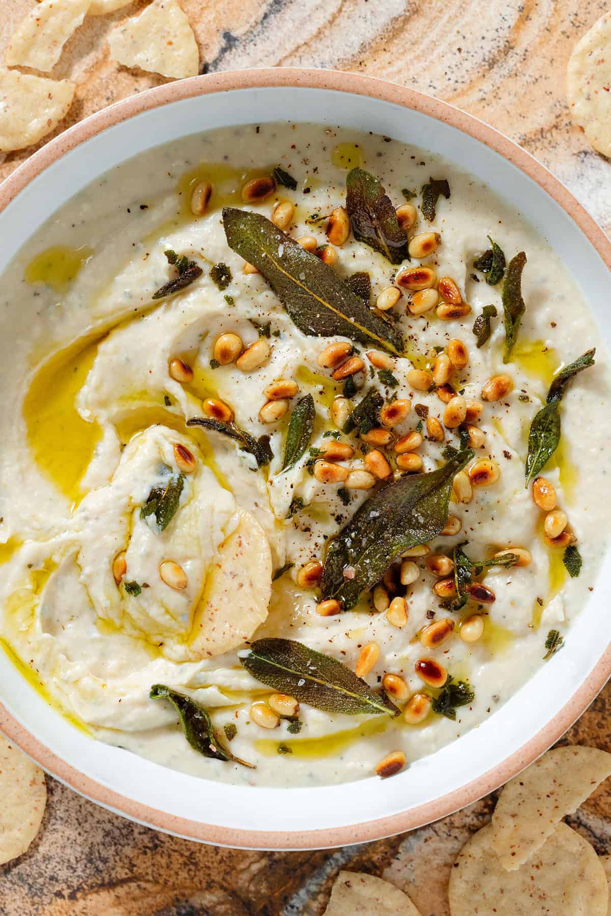 White bean dip in a low white bowl topped with fried sage leaves, toasted pine nuts, and olive oil with a round cracker dipped into it in the middle scooping the dip.
