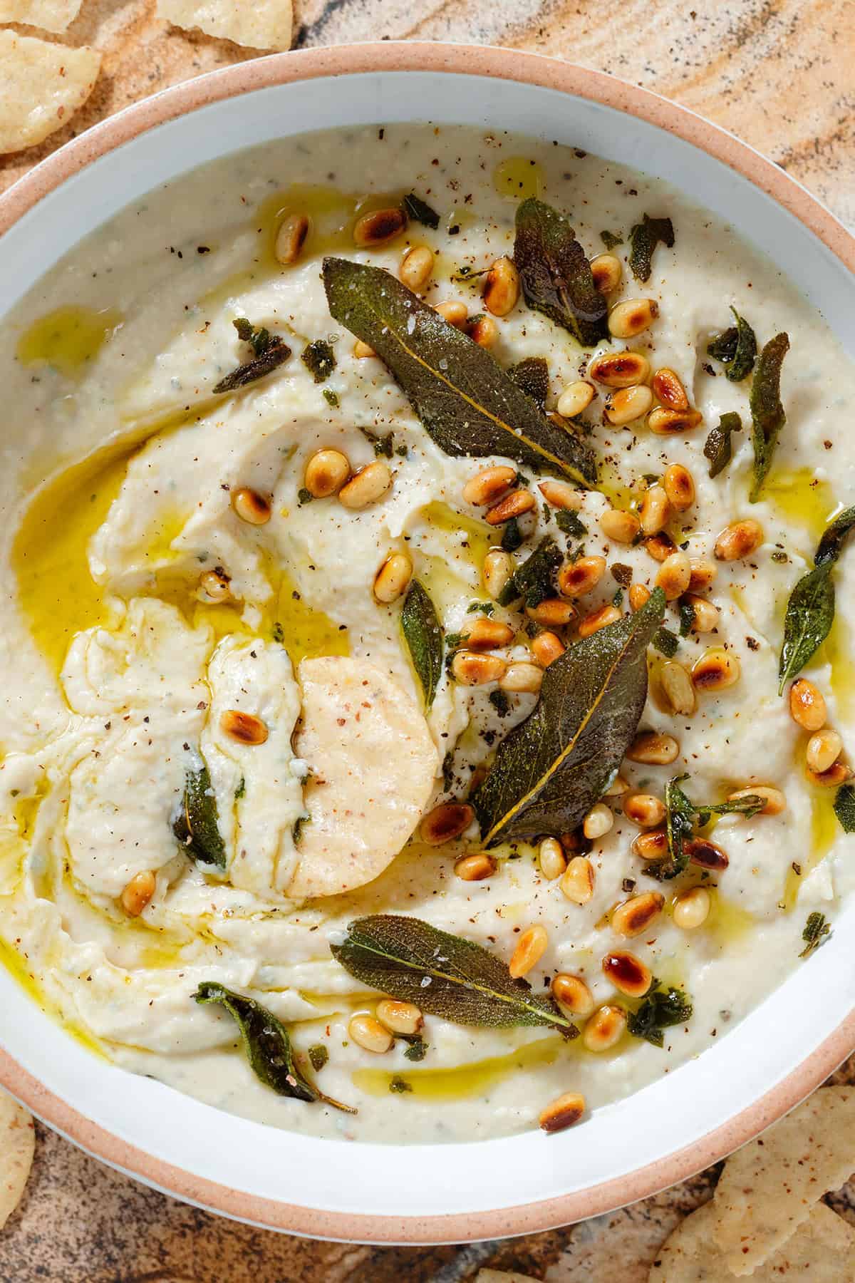 White bean dip in a low white bowl topped with fried sage leaves, toasted pine nuts, and olive oil with a round cracker dipped into it in the middle.