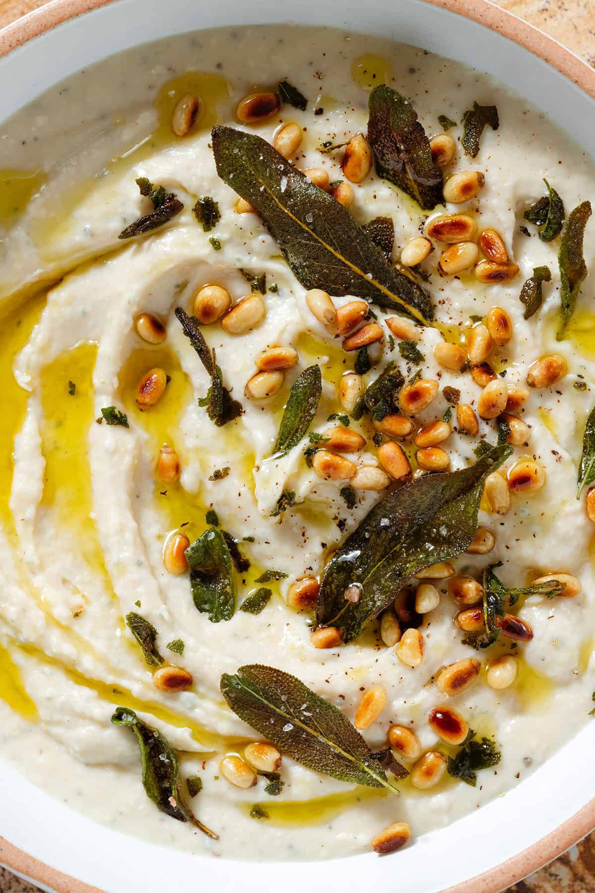 White bean dip in a low white bowl topped with fried sage leaves, toasted pine nuts, and olive oil, sprinkled with sumac.
