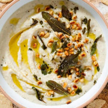 White bean dip in a low white bowl topped with fried sage leaves, toasted pine nuts, and olive oil.