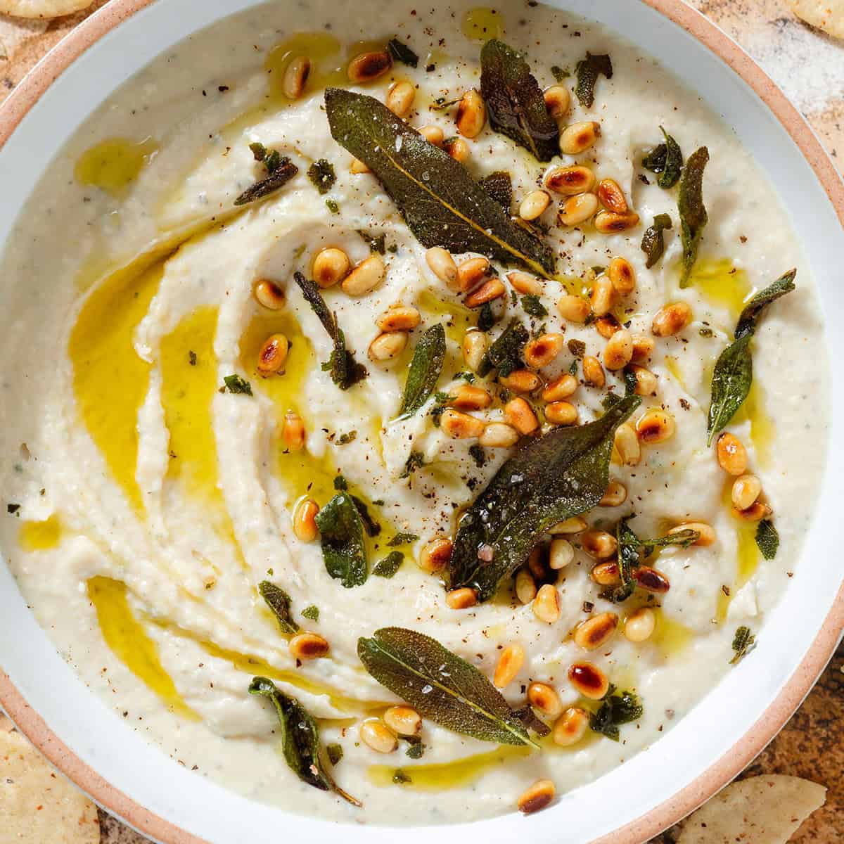 White bean dip in a low white bowl topped with fried sage leaves, toasted pine nuts, and olive oil.