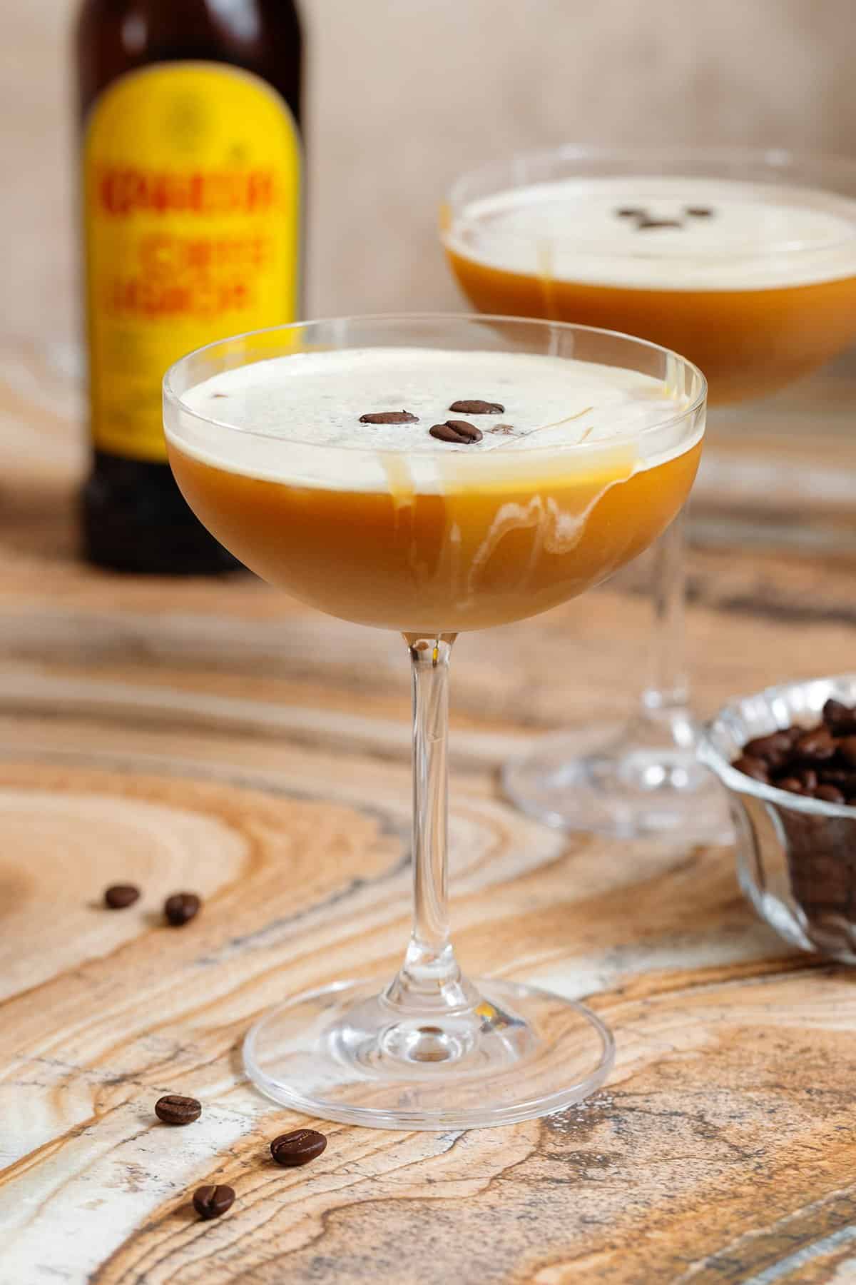A coupe glass with caramel espresso martini with thin foam garnished with 3 espresso beans and a drizzle of caramel sauce.