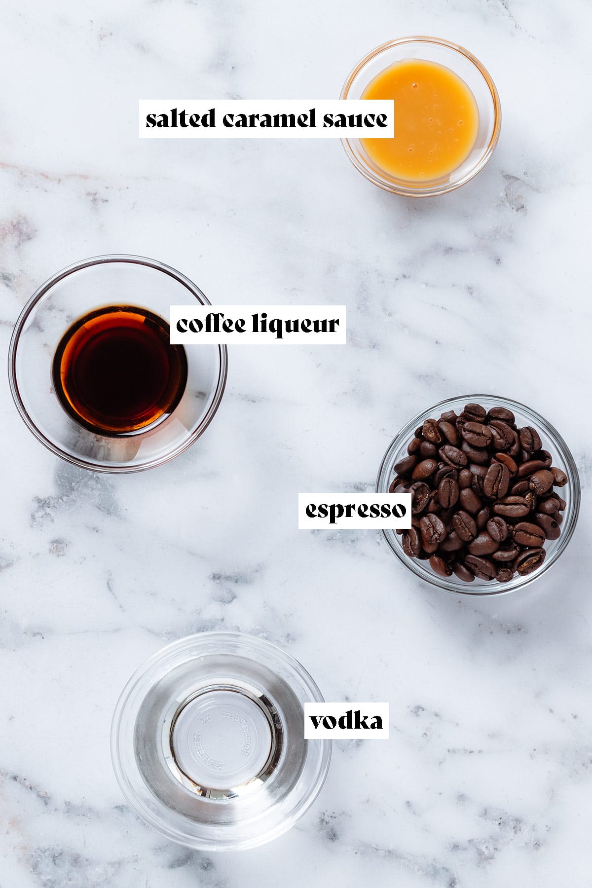Small glass bowls with espresso beans, coffee liqueur, vodka, and caramel sauce on marble background with text overlay.