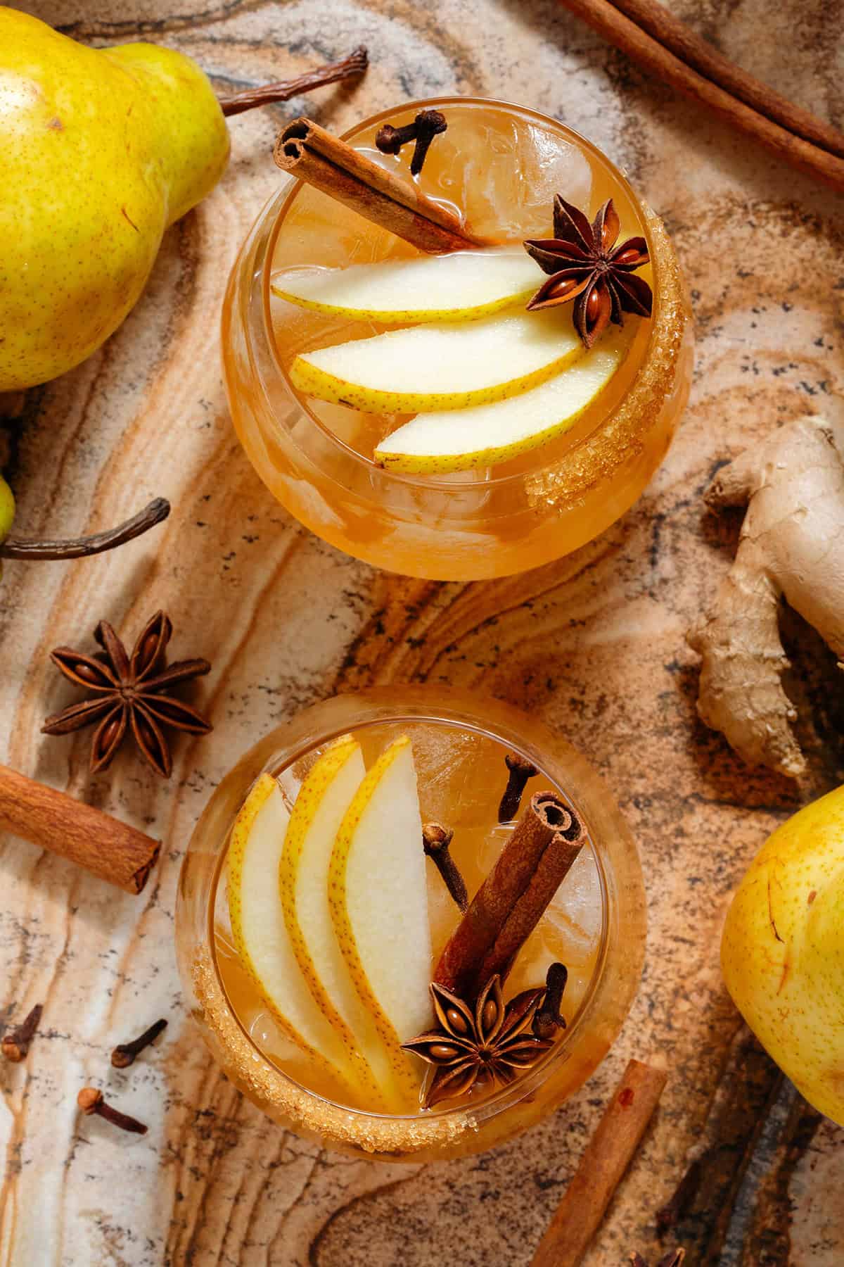 A deep orange pear cocktail in two short glasses with ice and a coarse sugar rim, garnished with sliced pear, a cinnamon stick, star anise, and cloves shot from above.