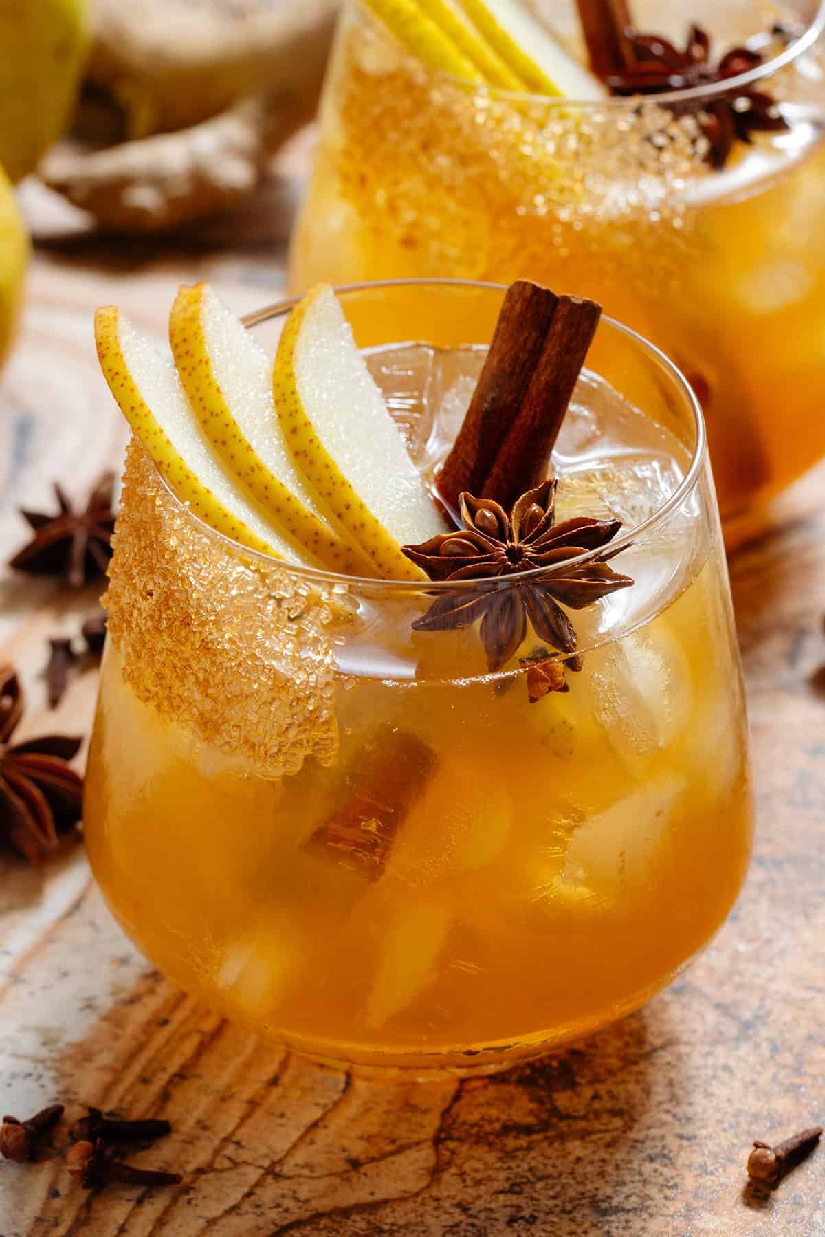 A deep orange pear cocktail in a short glass with ice and a coarse sugar rim, garnished with sliced pear, a cinnamon stick, star anise, and cloves.
