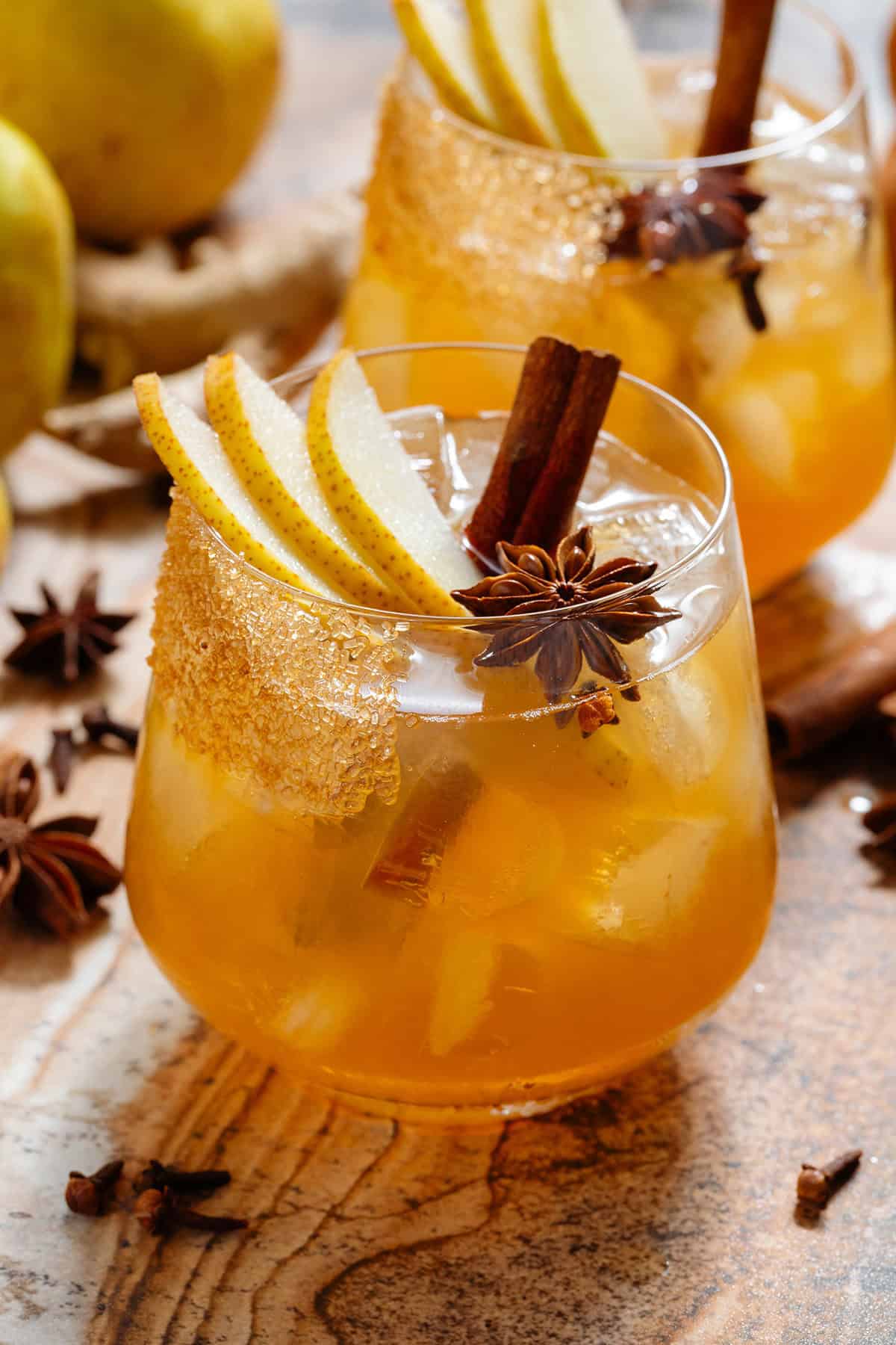 A deep orange pear cocktail in a short glass with ice and a coarse sugar rim, garnished with sliced pear, a cinnamon stick, star anise, and cloves.