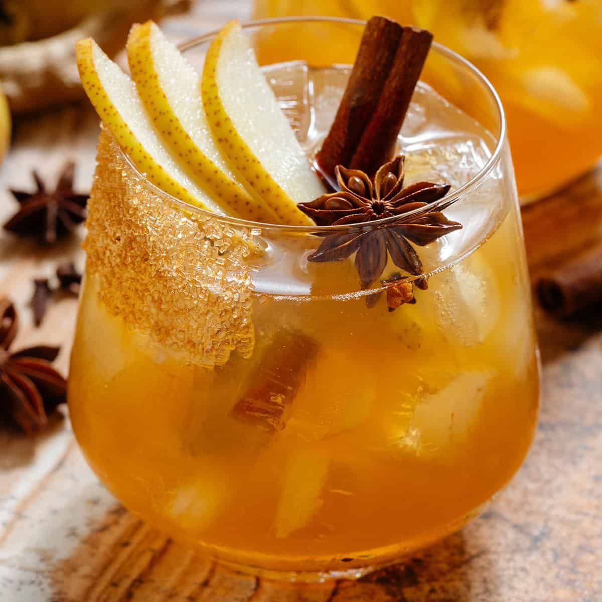 A deep orange pear cocktail in a short glass with ice and a coarse sugar rim, garnished with sliced pear, a cinnamon stick, star anise, and cloves.