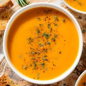 A white bowl with creamy butternut squash soup sprinkled with fresh thyme leaves and black pepper with toasted bread around it.