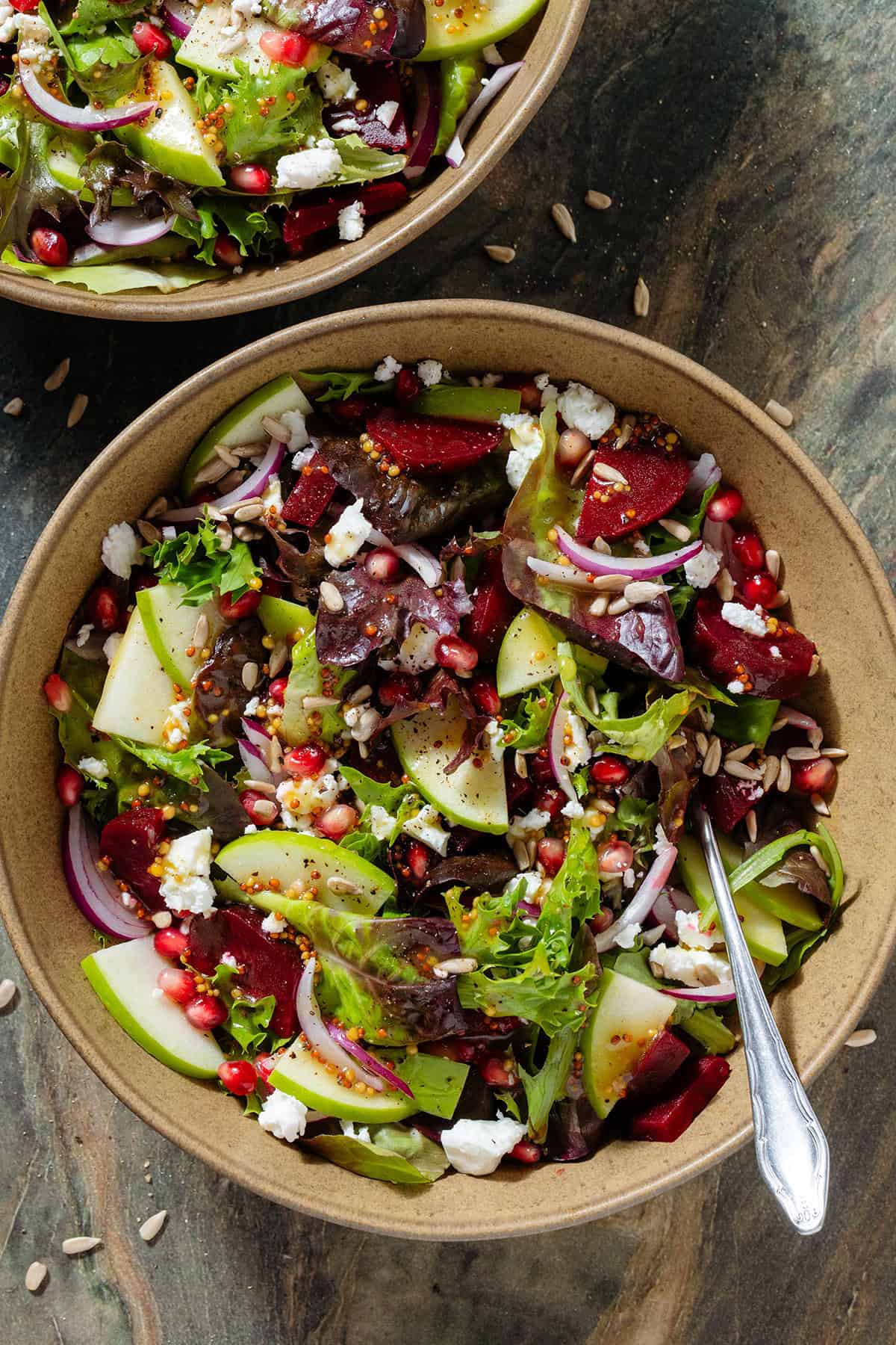 Salad with steamed beets, sliced apple, goat cheese, pomegranate arils, goat cheese, and whole grain mustard vinaigrette in a brown bowl with a fork inserted on the right.