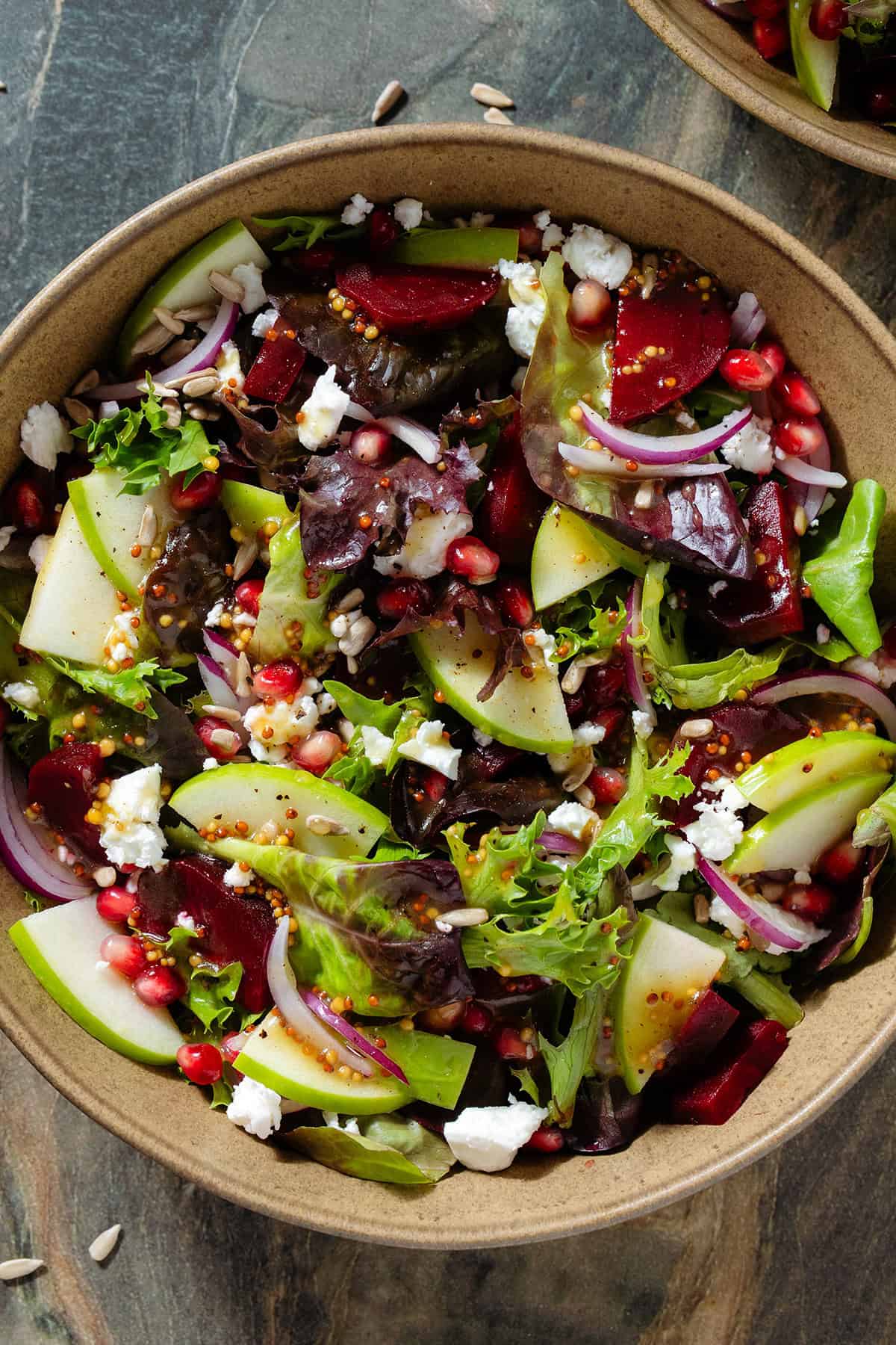 Salad with steamed beets, sliced apple, goat cheese, pomegranate arils, and whole grain mustard vinaigrette in a brown bowl.