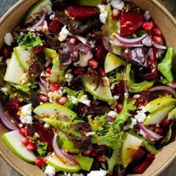 Salad with steamed beets, sliced apple, goat cheese, pomegranate arils, goat cheese, and whole grain mustard vinaigrette in a brown bowl.