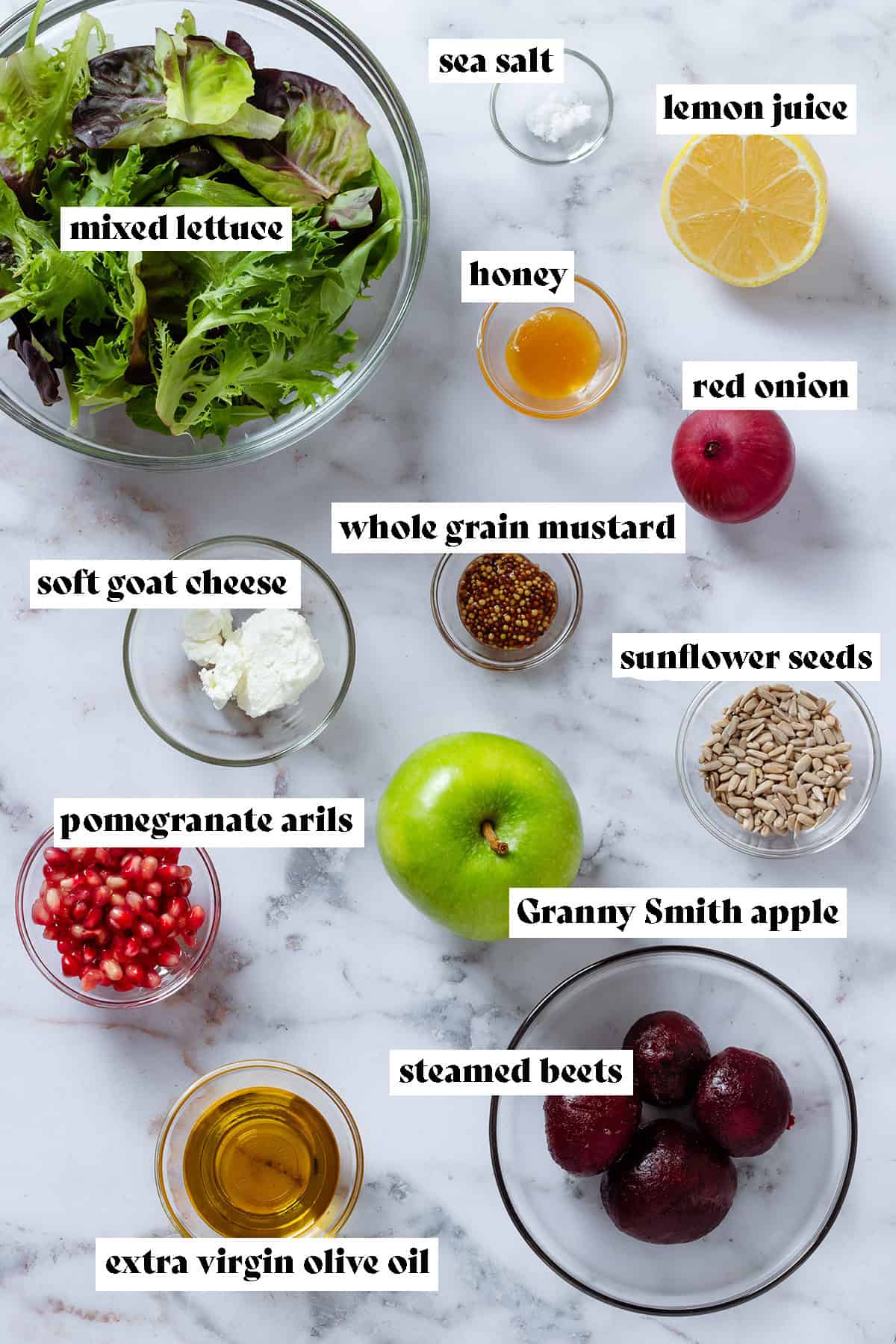 Ingredients like lettuce leaves, pomegranate arils, steamed beets, sunflower seeds, olive oil, whole grain mustard, goat cheese, and more in glass bowls.