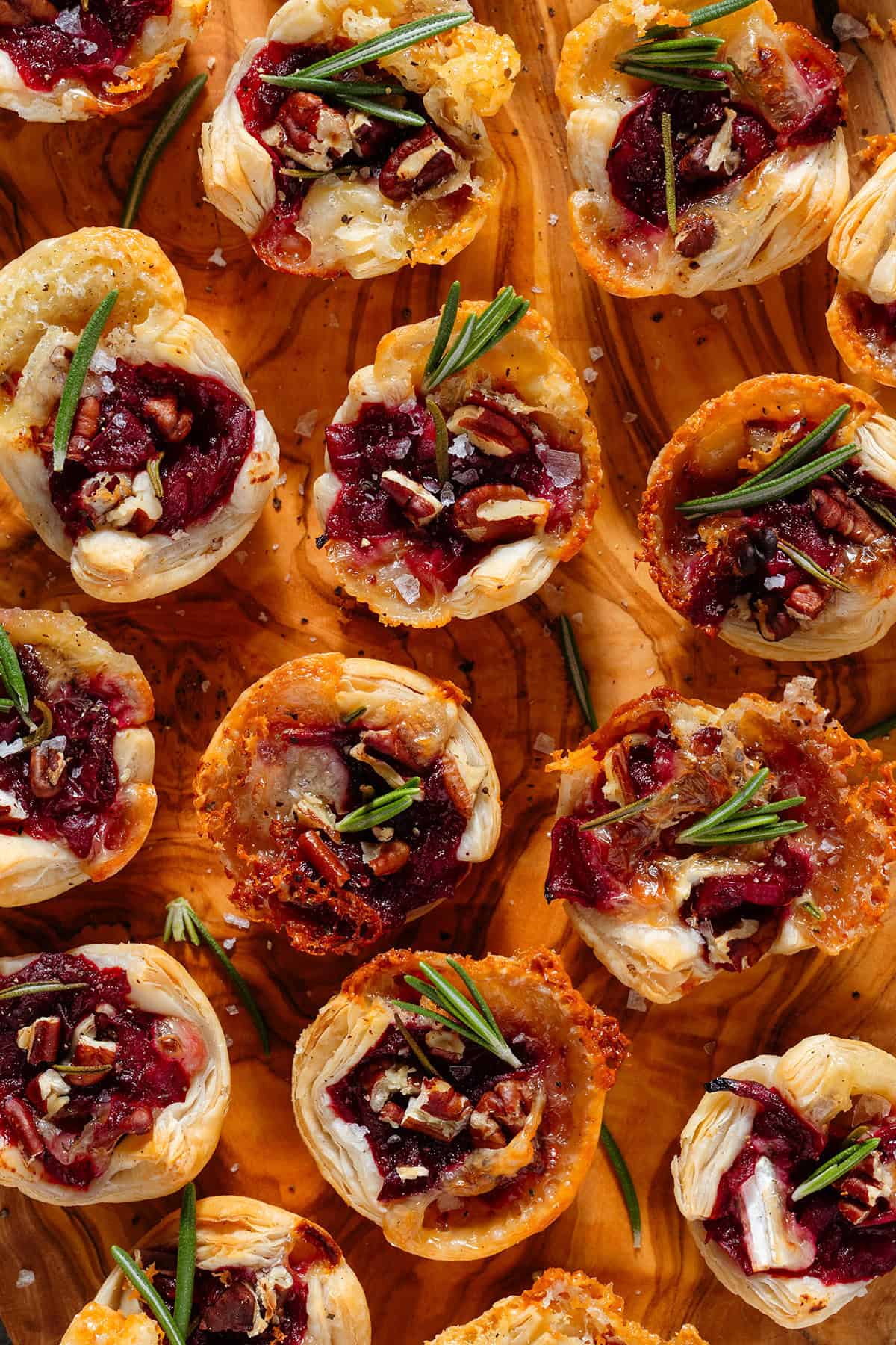 Small puff pastry cups with melted brie, cranberry sauce, pecans, and fresh rosemary sprinkled with flaky salt and pepper on a wooden serving board.