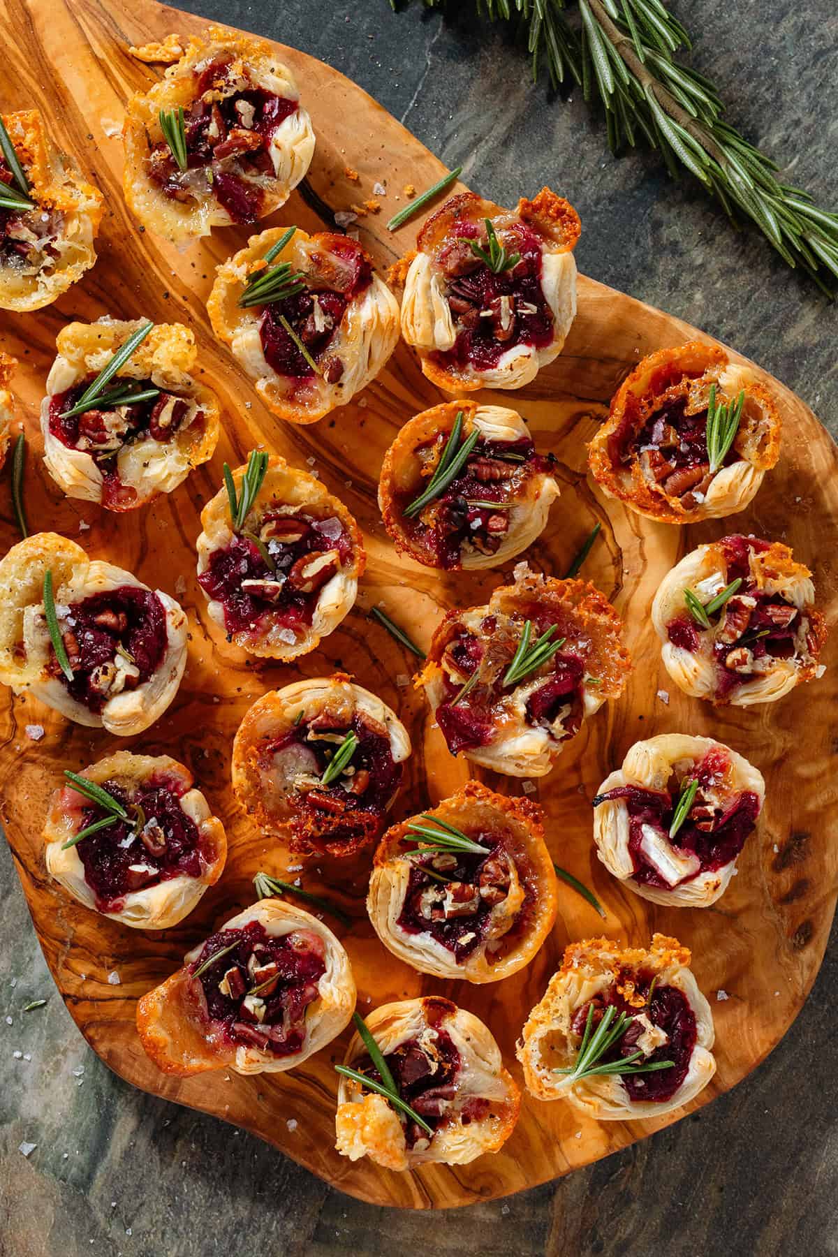 Small puff pastry cups with melted brie, cranberry sauce, pecans, and fresh rosemary sprinkled with sea salt and pepper on a wooden serving board.