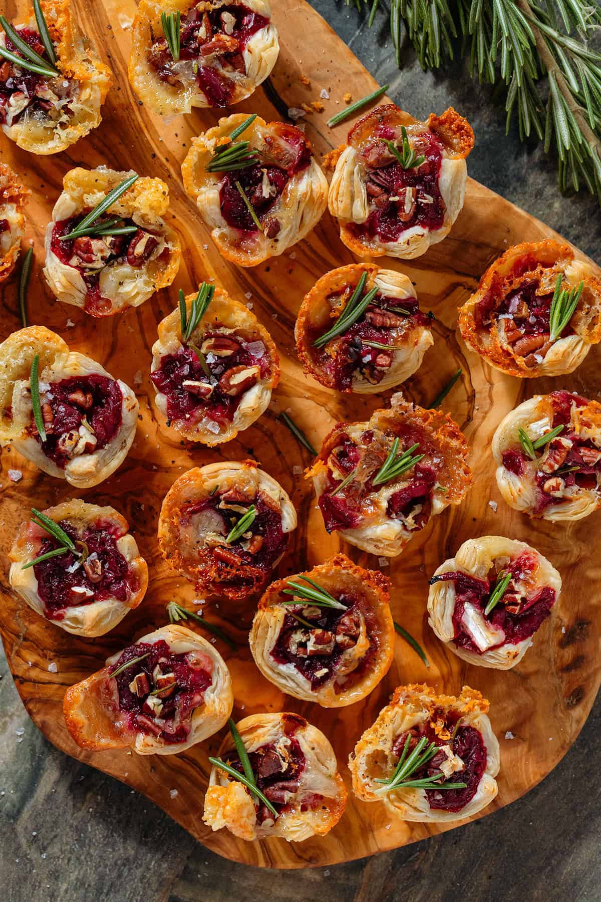 Small puff pastry cups with melted brie, cranberry sauce, pecans, and fresh rosemary sprinkled with sea salt and pepper on a wooden serving board.