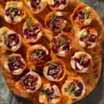 Small puff pastry cups with melted brie, cranberry sauce, pecans, and fresh rosemary sprinkled with sea salt and pepper on a wooden serving board.