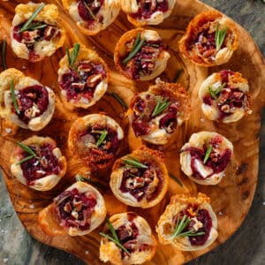 Small puff pastry cups with melted brie, cranberry sauce, pecans, and fresh rosemary sprinkled with sea salt and pepper on a wooden serving board.