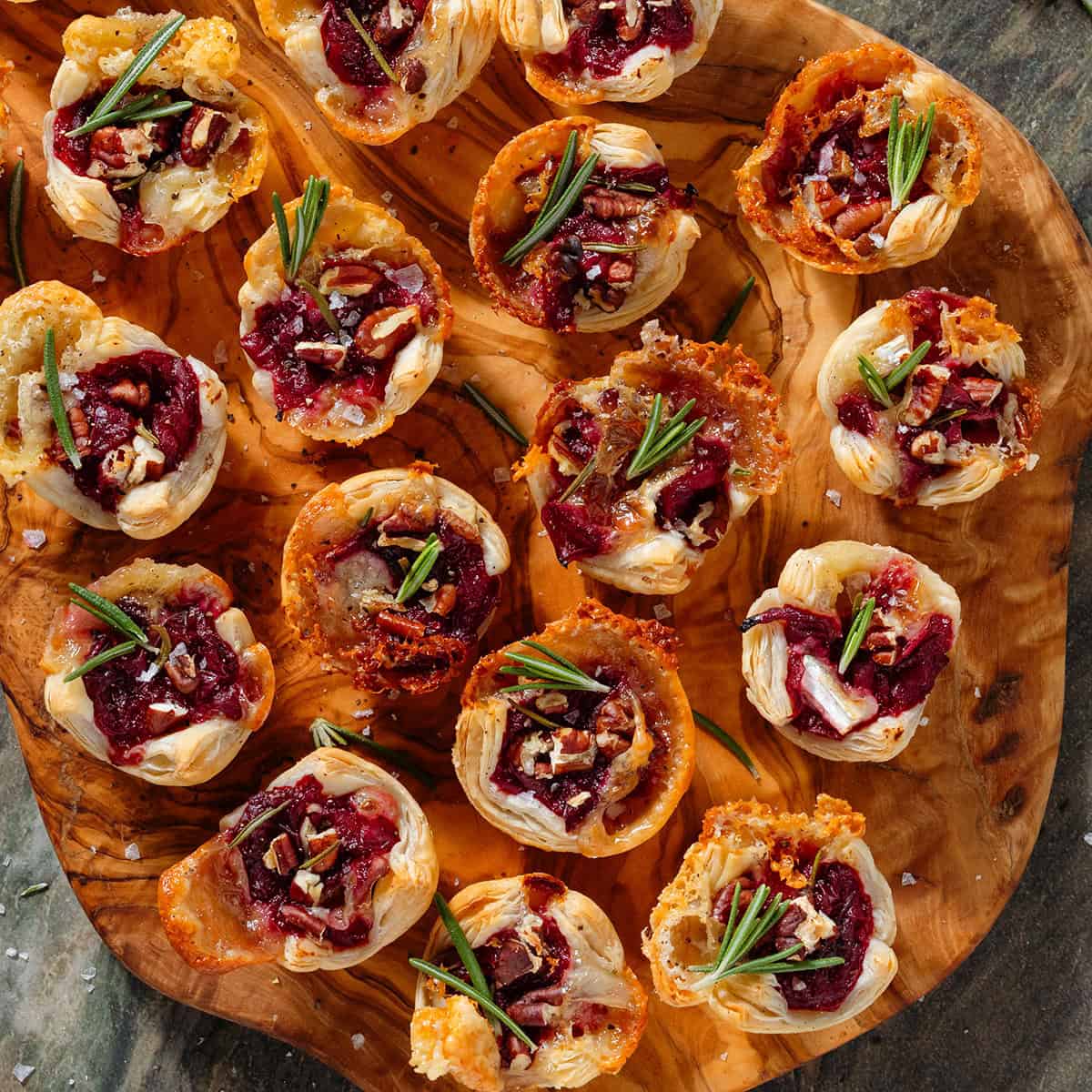 Small puff pastry cups with melted brie, cranberry sauce, pecans, and fresh rosemary sprinkled with sea salt and pepper on a wooden serving board.