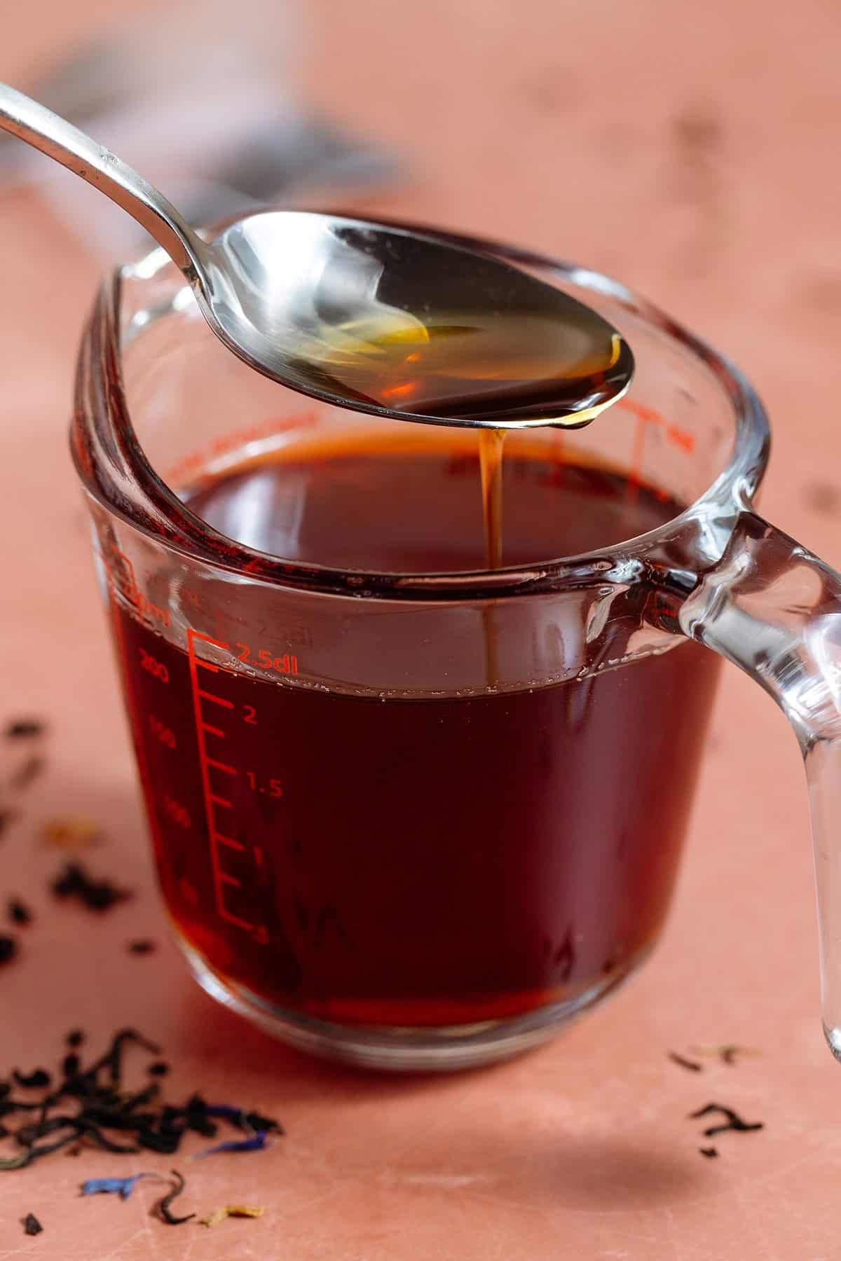 Dark brown earl grey syrup in a large glass measuring cup with a spoon picking up some of the syrup.