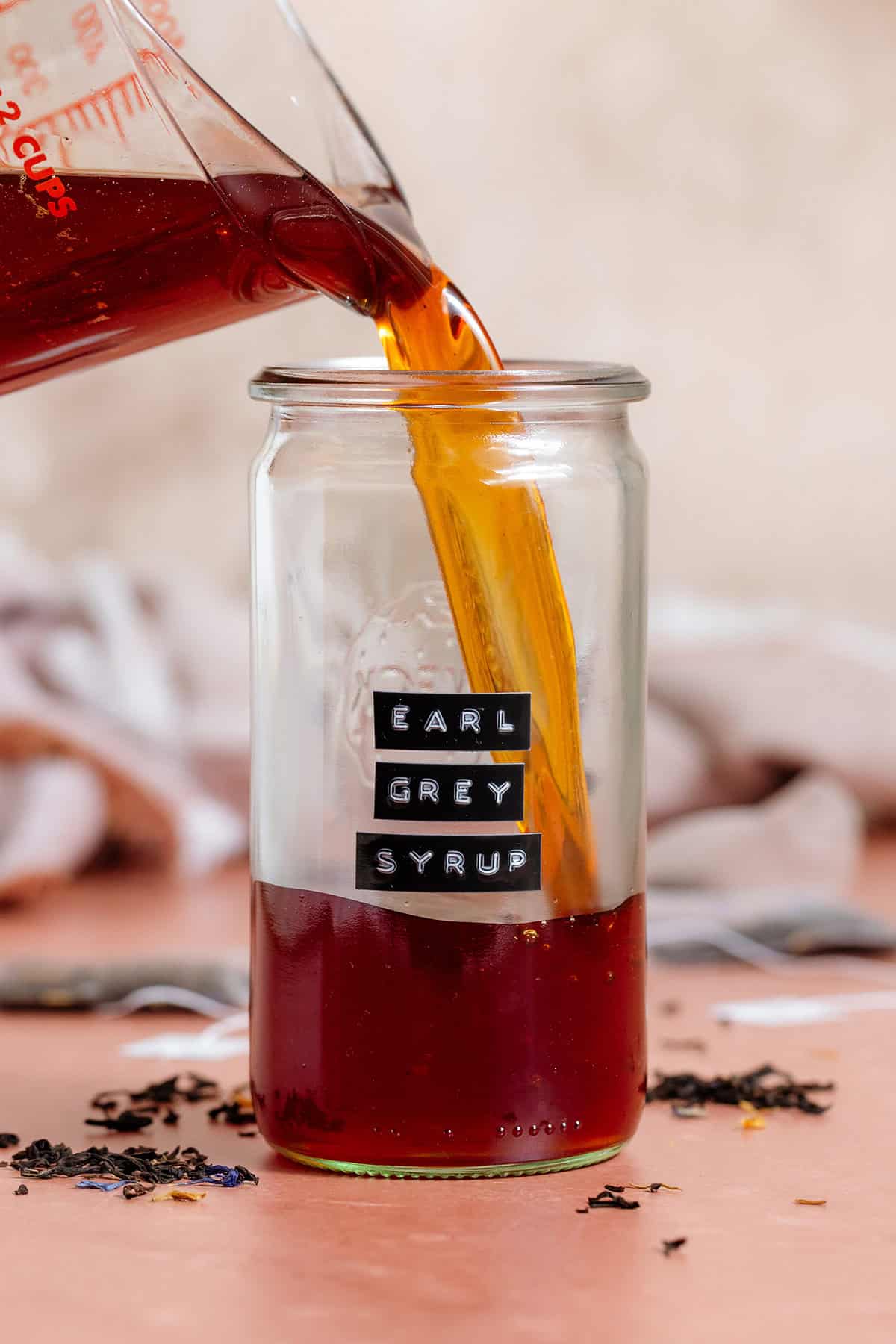 Dark brown syrup being poured into a tall glass jar with an embossed label that says "earl grey syrup".