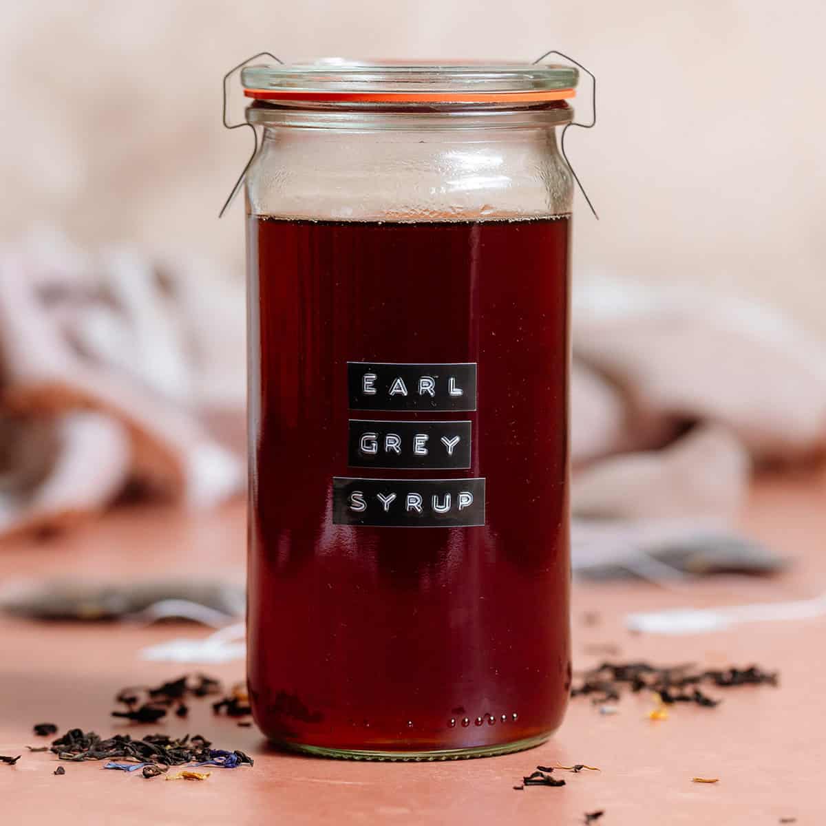 A tall glass jar with earl grey syrup closed with a lid with a black embossed label on the front of the jar.