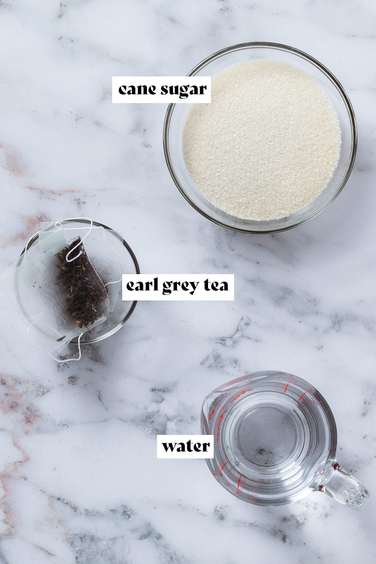 Glass bowls with cane sugar, earl grey teabags, and water on a marble background with text overlay.
