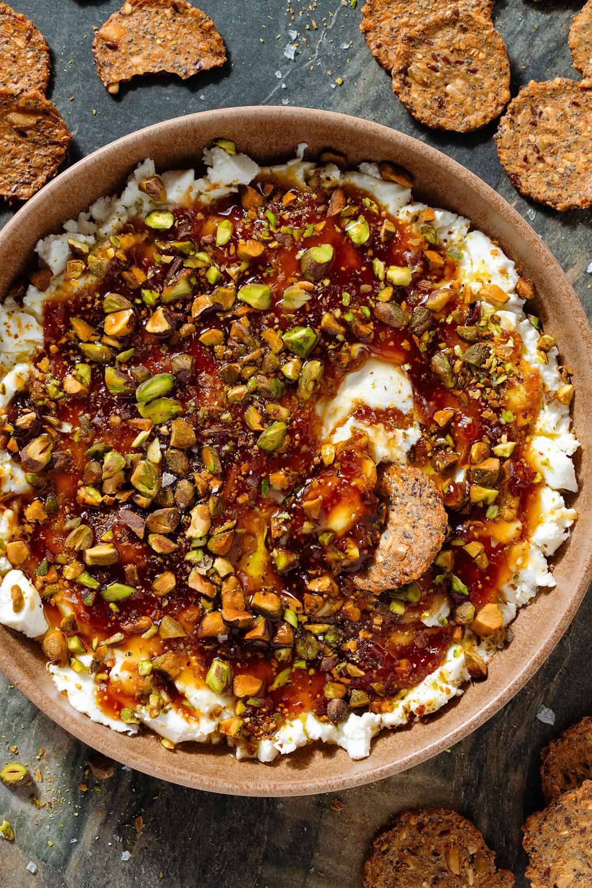 Soft goat cheese topped with fig jam, olive oil, and chopped roasted pistachios in a low brown bowl with a cracker scooping up the dip.