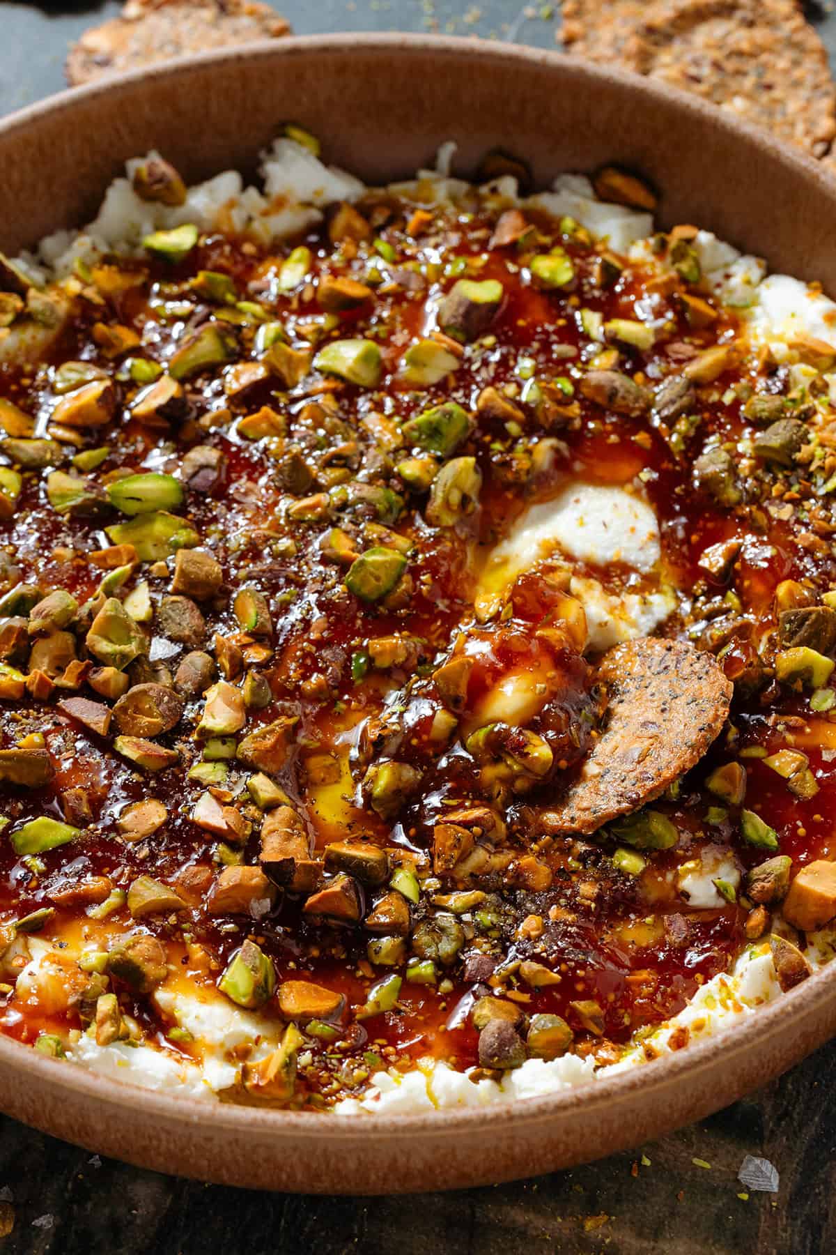 Soft goat cheese topped with fig jam, olive oil, honey, and chopped roasted pistachios in a low brown bowl with a cracker scooping up the dip.