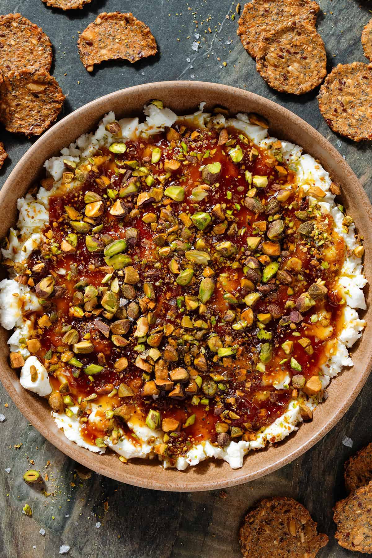 Soft goat cheese topped with fig jam, olive oil, and chopped roasted pistachios in a low brown bowl.
