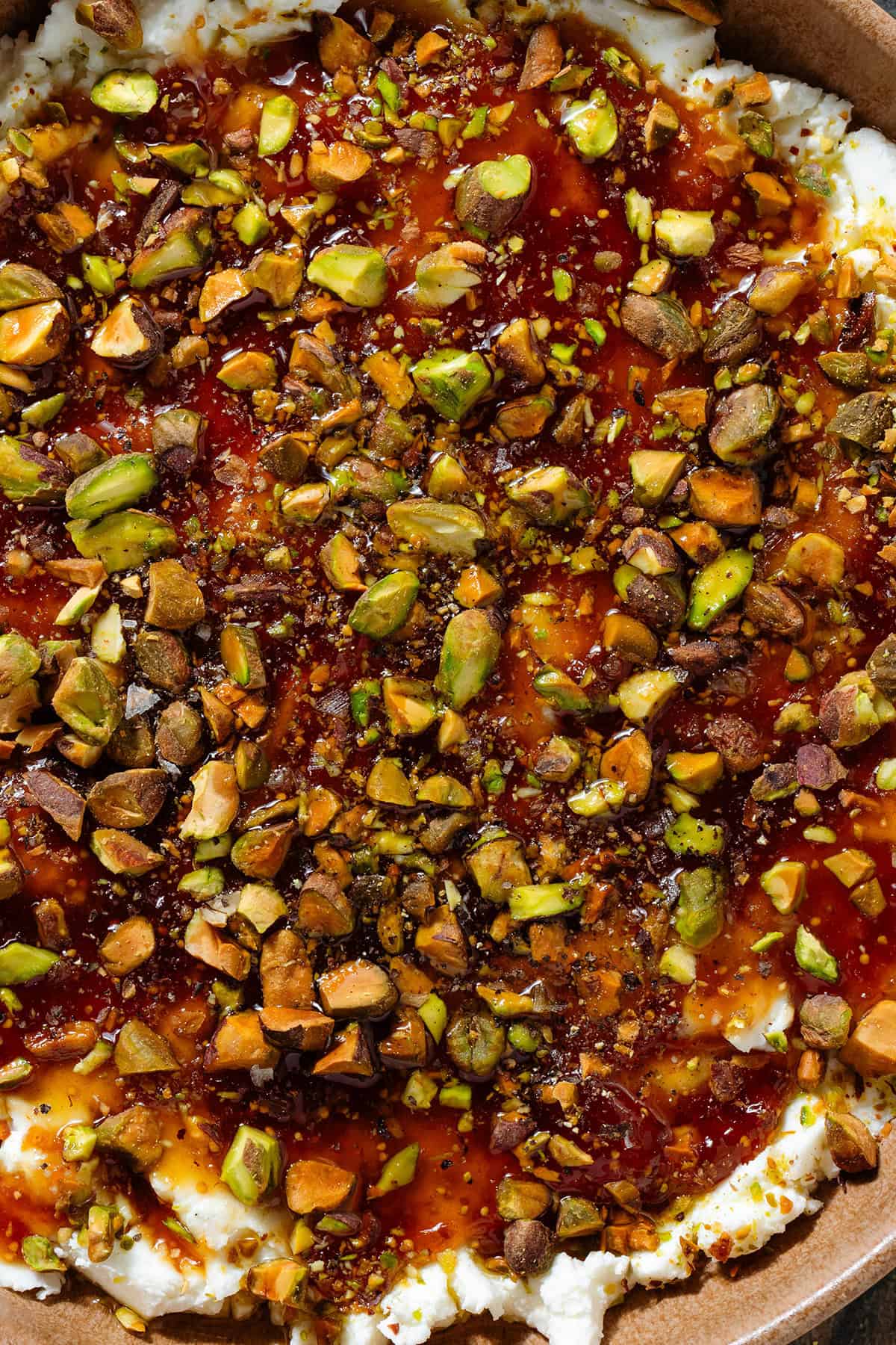 Soft goat cheese topped with fig jam, olive oil, honey, flaky sea salt, and chopped roasted pistachios on a brown plate.