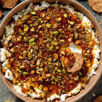 Soft goat cheese topped with fig jam, olive oil, and chopped roasted pistachios in a low brown bowl with a cracker scooping up the dip.