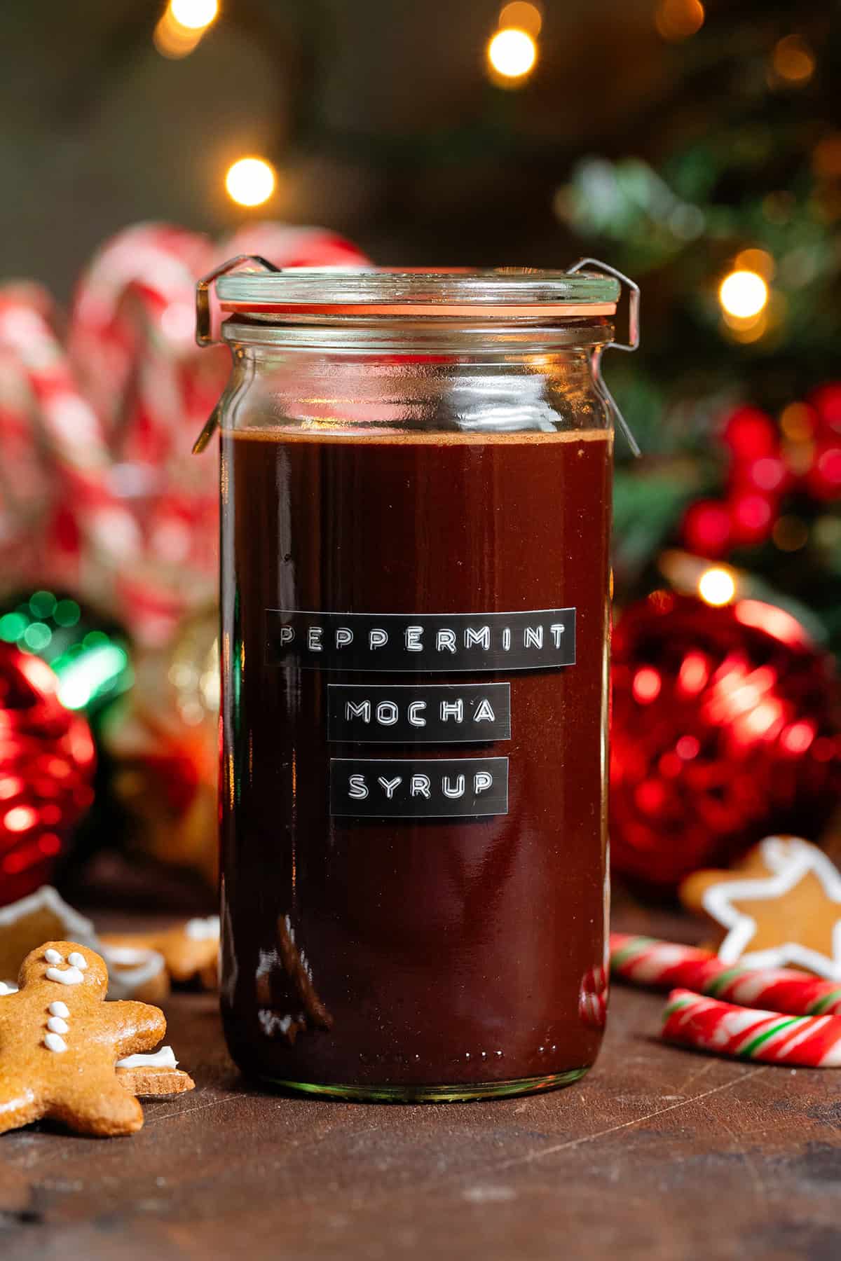 A glass jar with homemade chocolate peppermint syrup with a black embossed label on the front with gingerbread cookies and candy canes in the background.