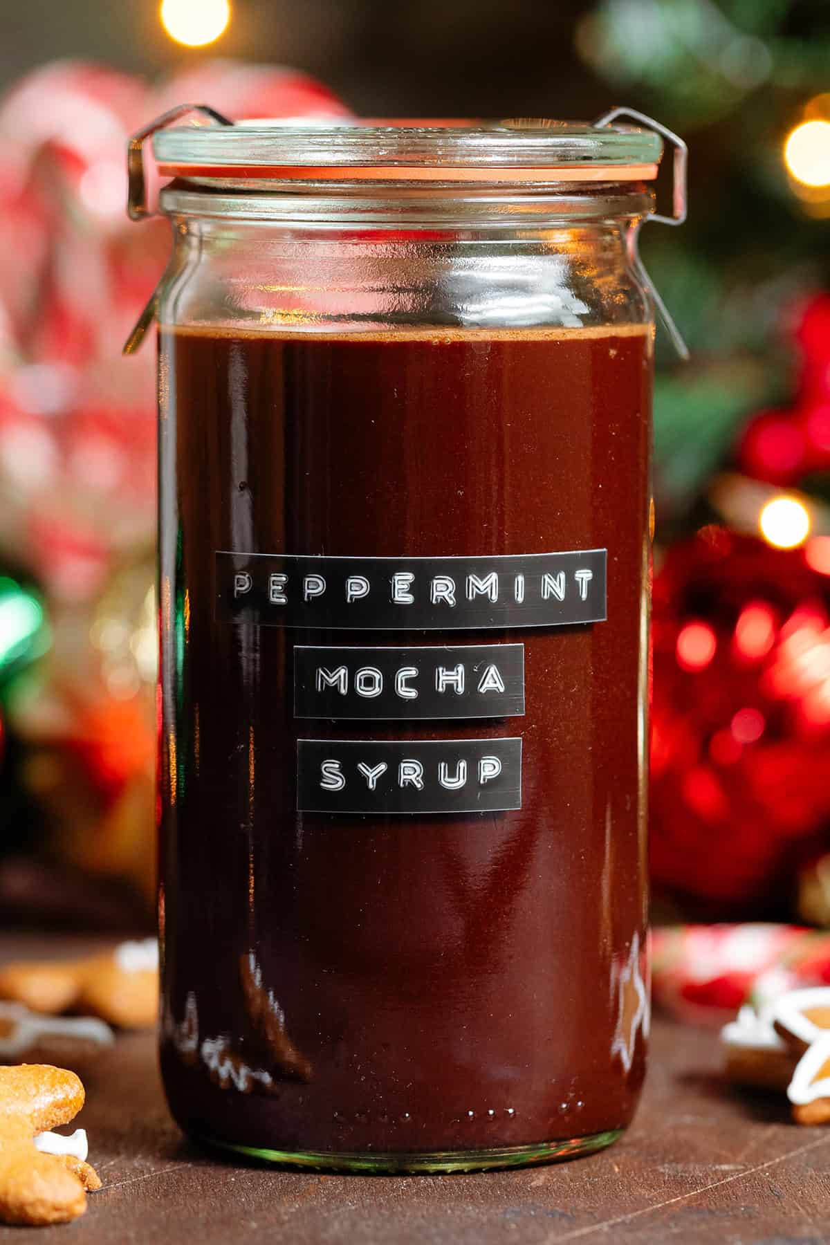 A glass jar with homemade chocolate peppermint syrup with a black embossed label on the front with candy canes in the background.