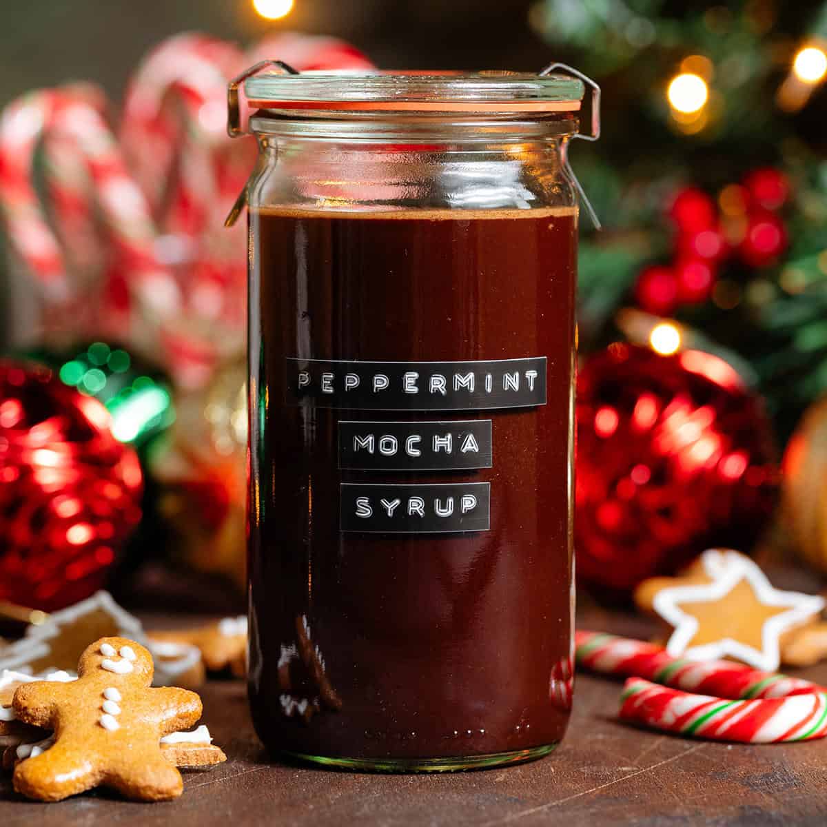 A glass jar with homemade chocolate peppermint syrup with a black embossed label on the front with gingerbread cookies and peppermint candy canes in the background.