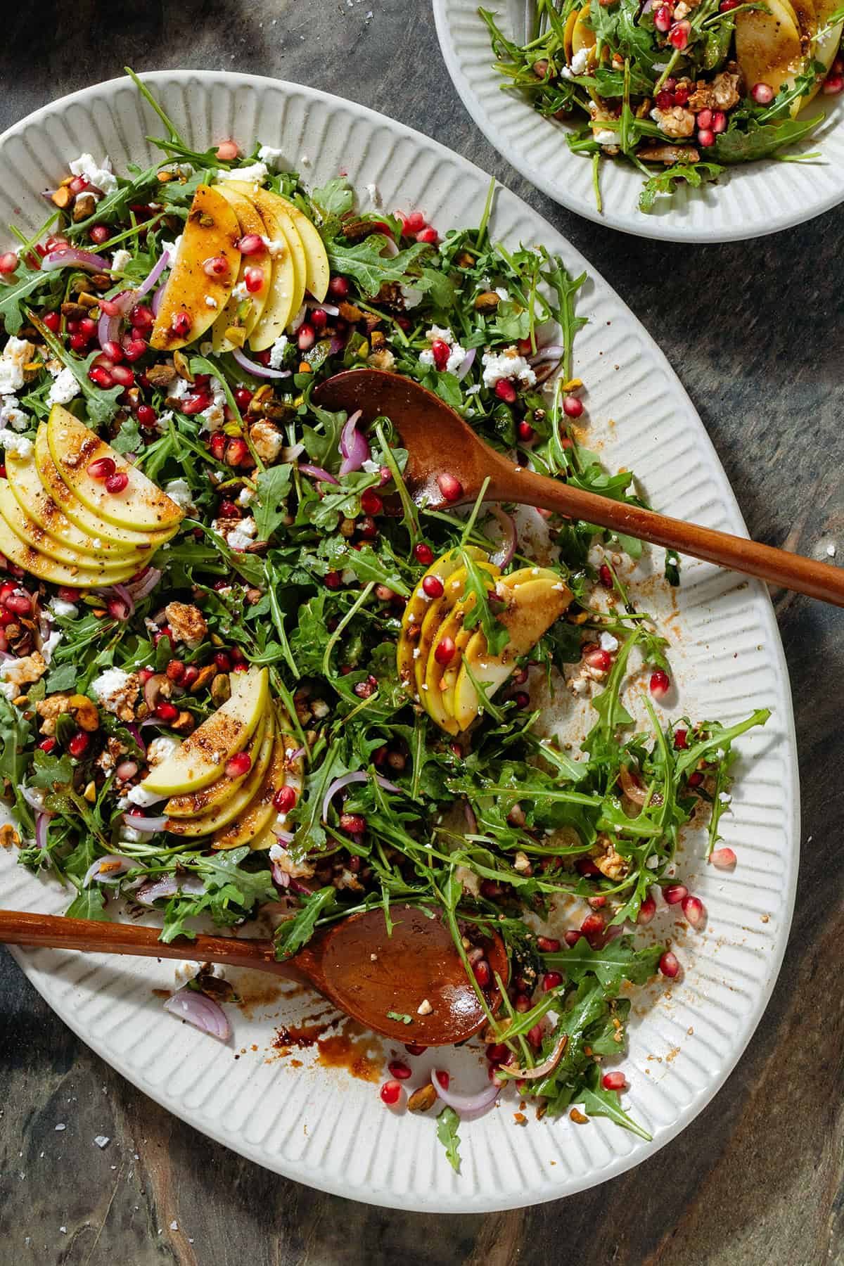 Arugula salad with sliced apples and pomegranate arils on a large serving platter with wooden serving spoons and in a small bowl in the top right.