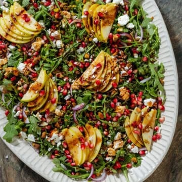 Arugula salad with sliced apples, pomegranate arils, feta, and red onion on a large serving plate.