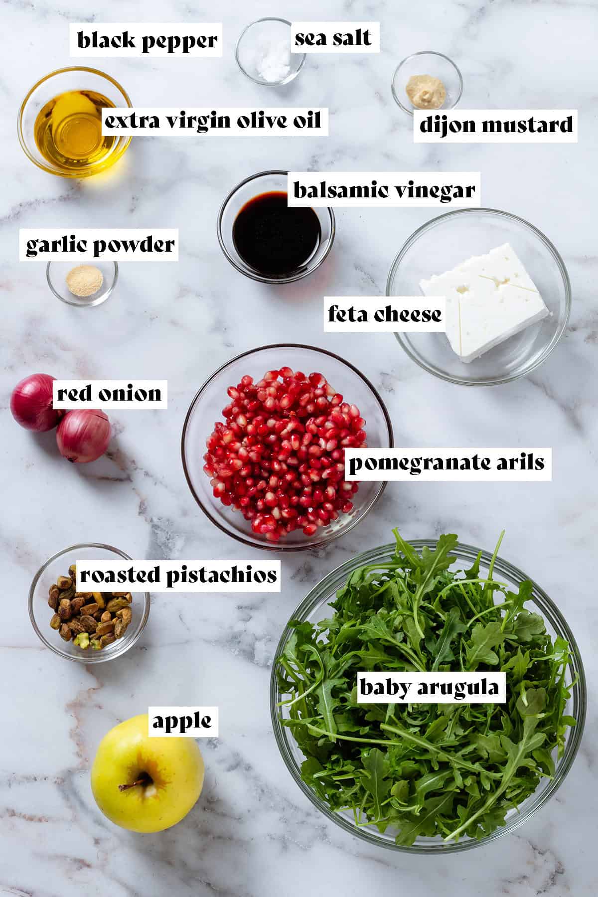 Ingredients like baby arugula, pomegranate arils, feta, pistachios, balsamic vinegar, olive oil, spices, and other ingredients in glass bowls.