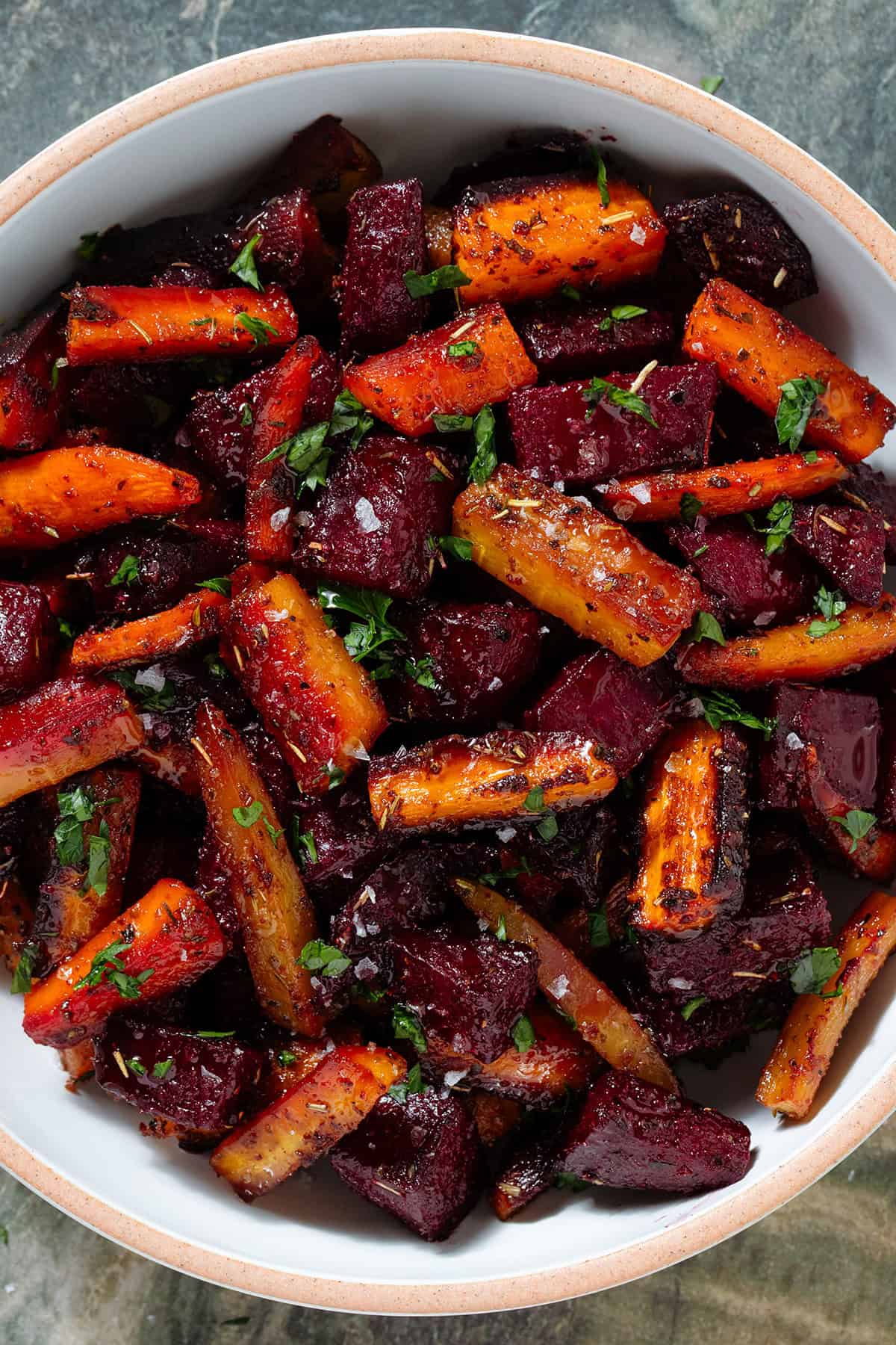 Caramelized roasted beets and carrots with balsamic vinaigrette garnished with chopped fresh parsley and flaky sea salt in a white bowl.